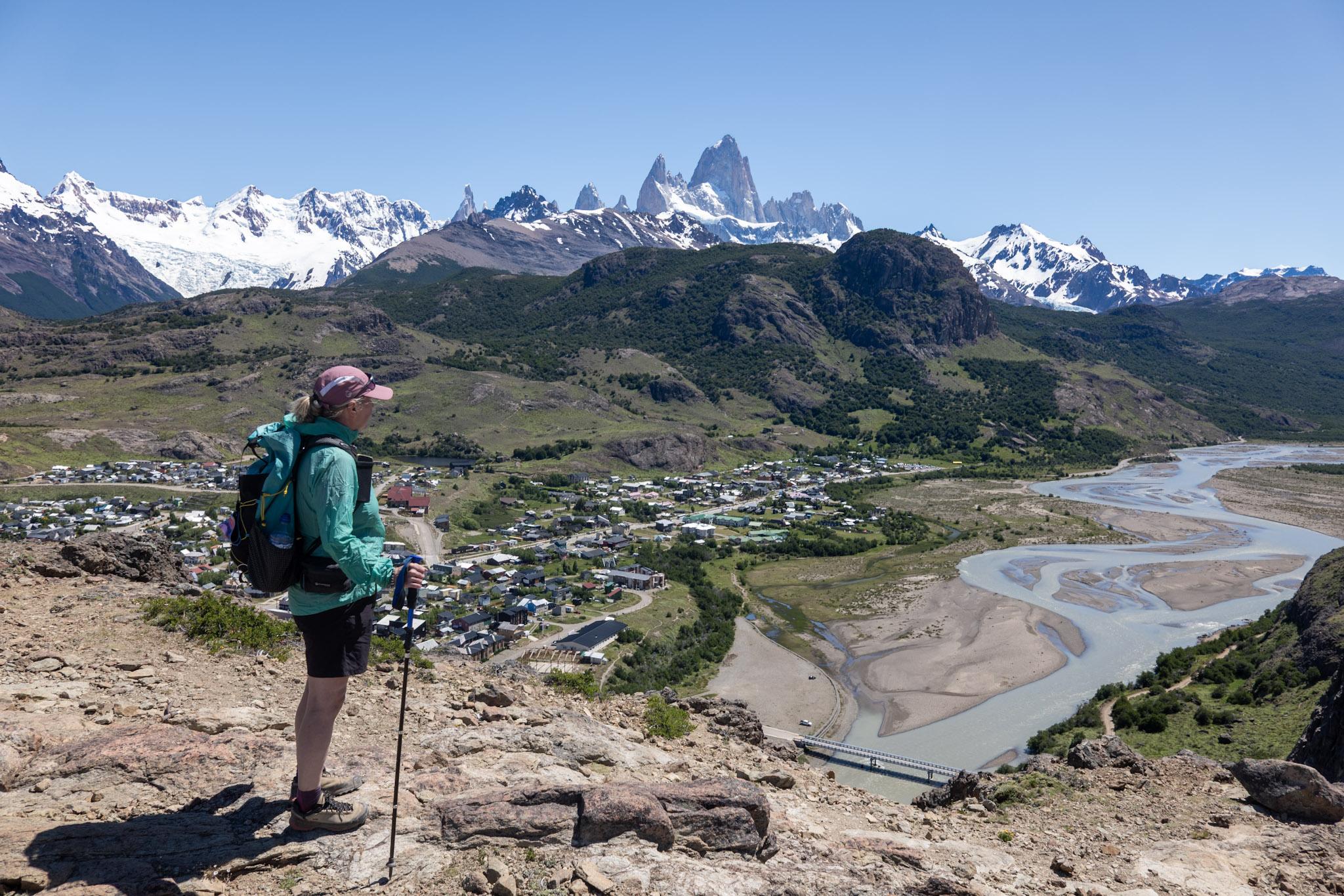 A person with light green jacket stands on a cliff edge. Beyond lies the town of El Chalten and beyond that the Cerro Torre and Fitzroy mountain ranges