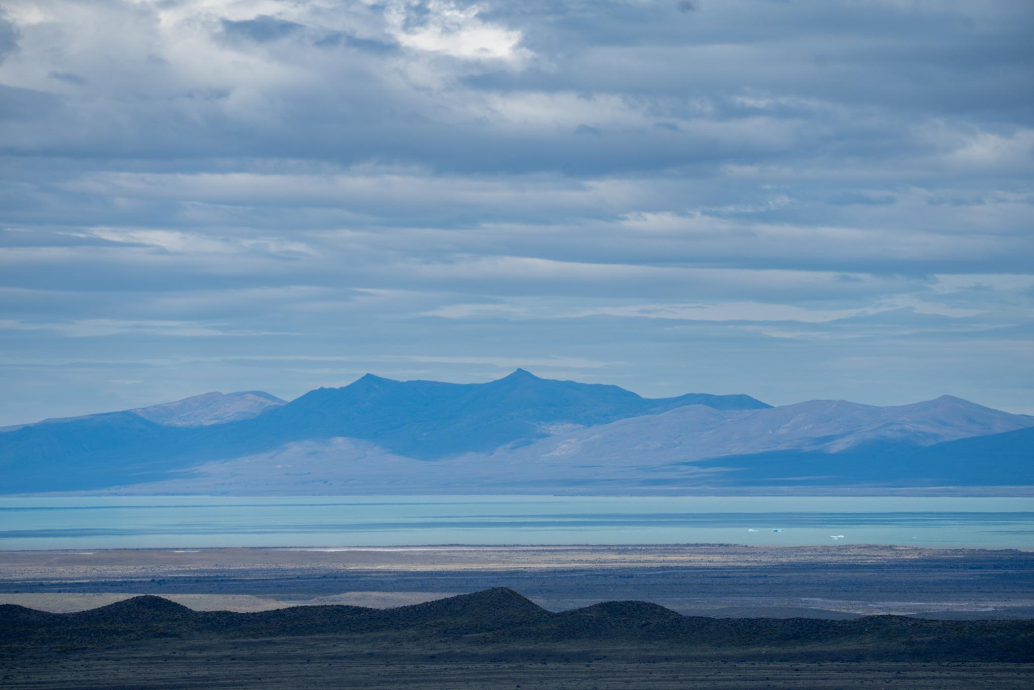 A series of horizontal lines from the close wavy hills in shadow to the plains leading to an icy blue lake. A couple of icebergs to the right. The outline of dark hills beyond with clouds above