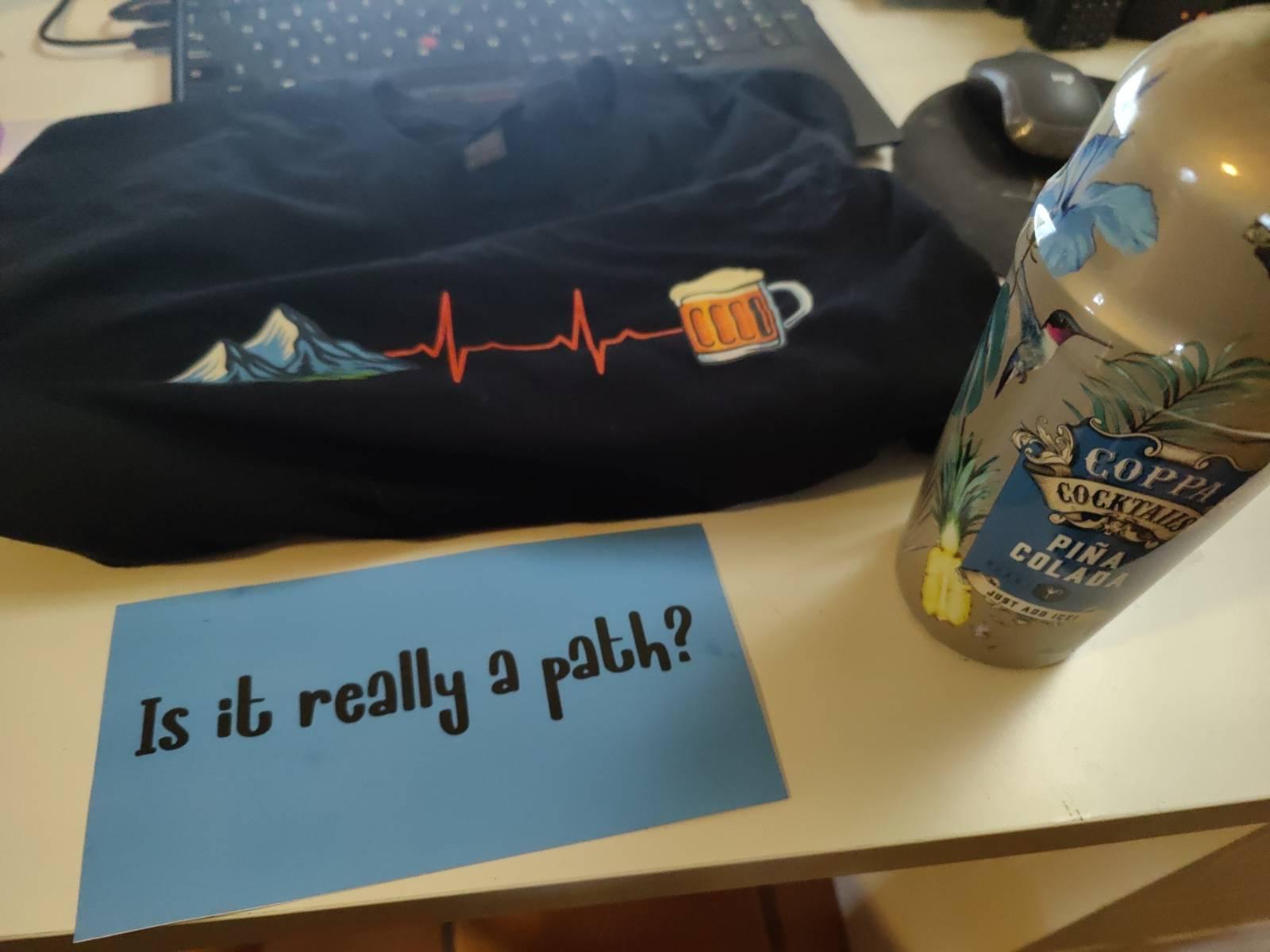 A tee shirt with mountains leading to beer emblem, a bottle of pina colada and a card that reads "Is it really a path?"