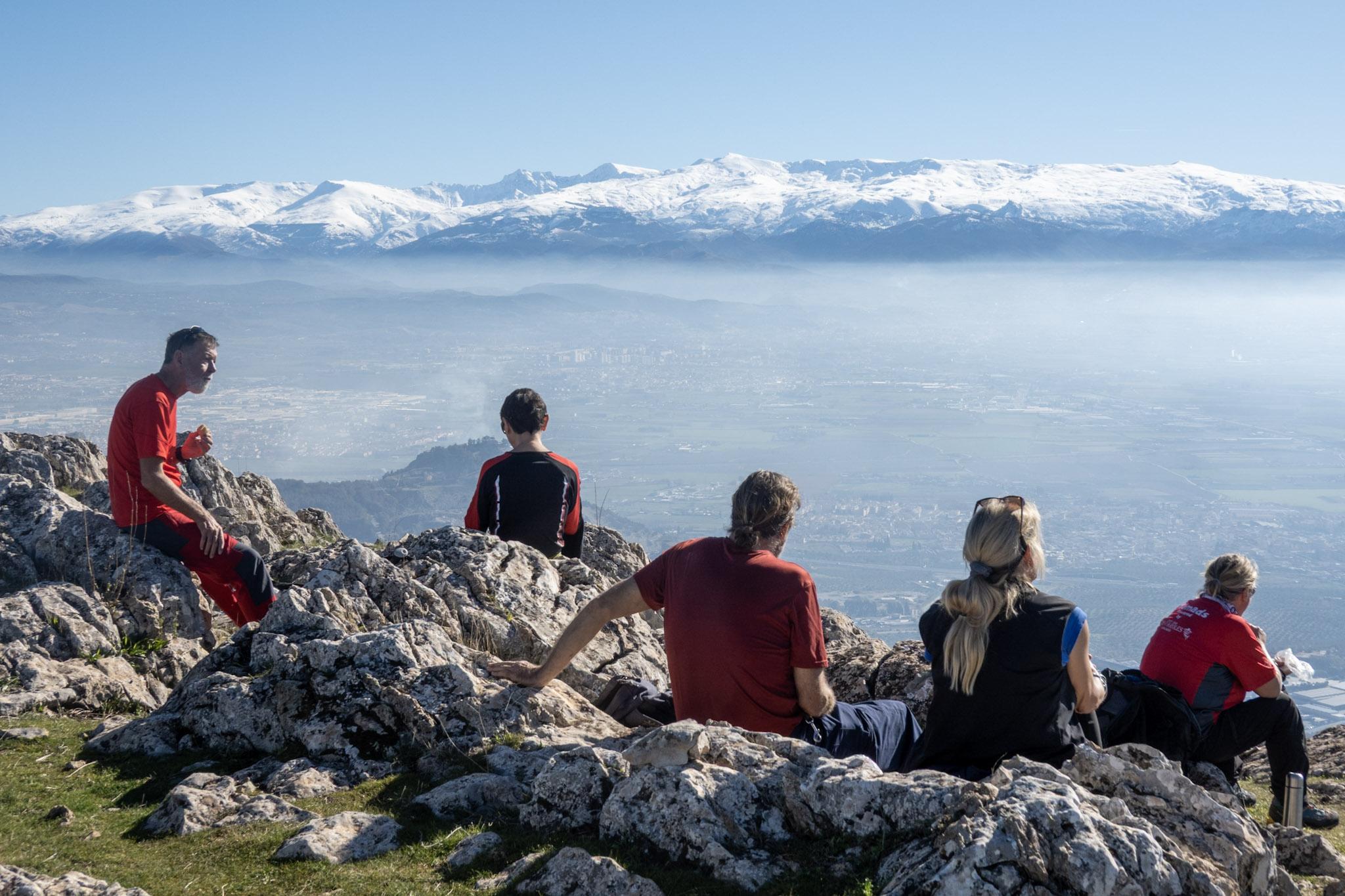 5 people sit at the top of a mountains eating lunch in bright clothing. beyond there is some low lying mist and then white mountains rising up above the mists
