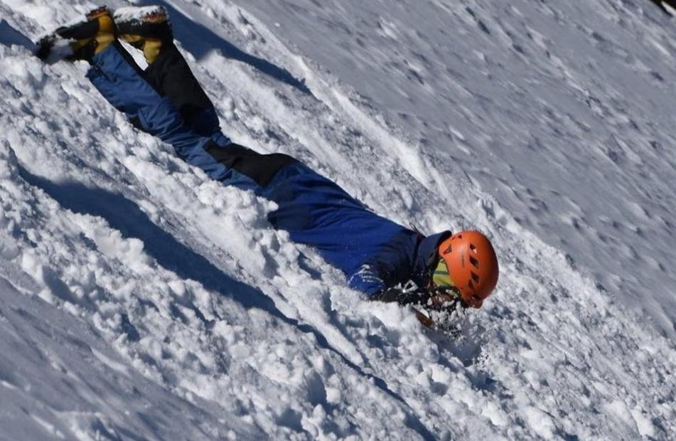 I'm heading down a steep snow slope head first. I am dressed in blue with orange helmet. I will stop by turning myself around and braking using an ice axe