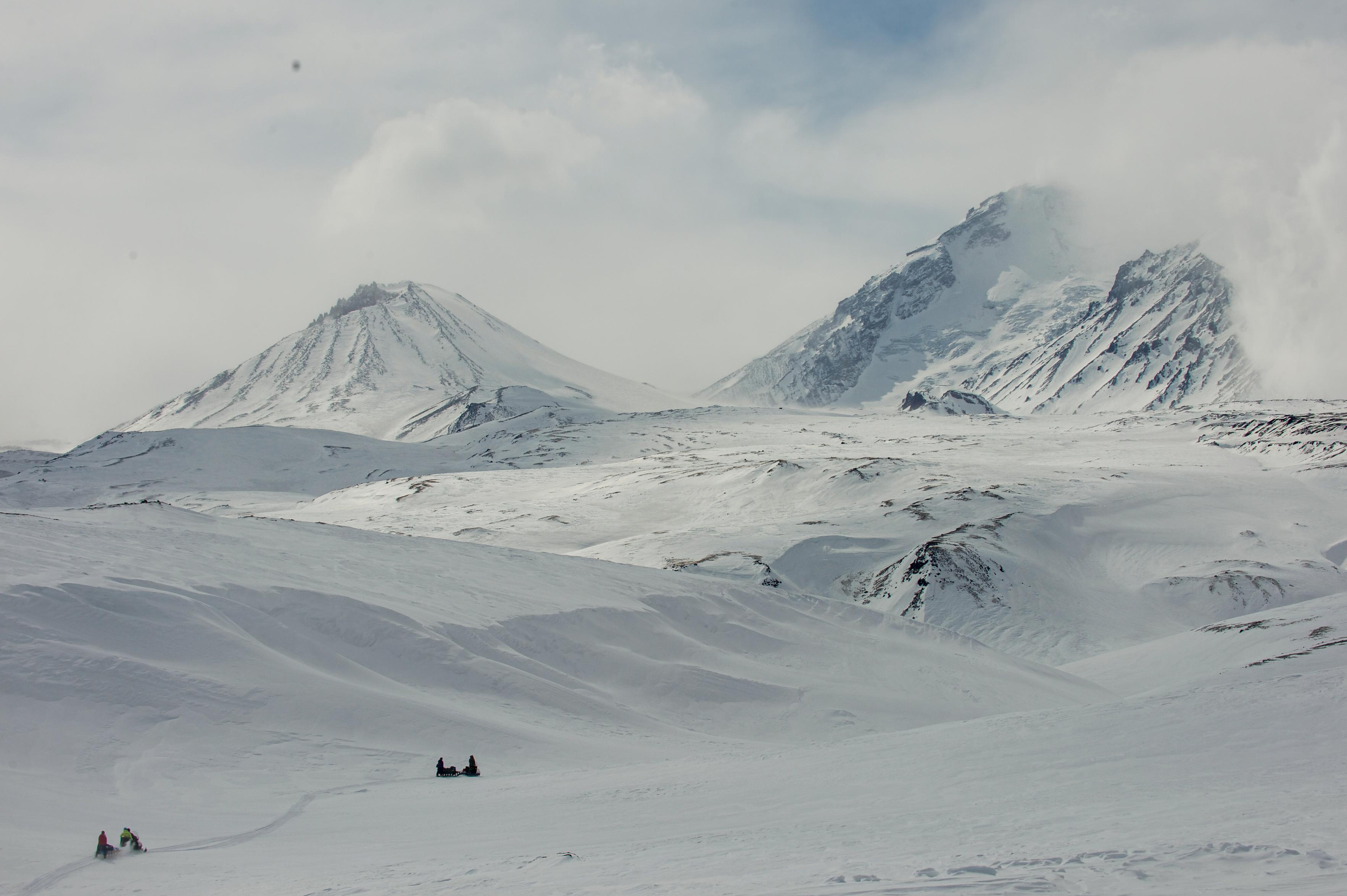 Two snowmobiles and sleds in the bottom left navigate a show covered terrain. Outline of higher mountains in the background
