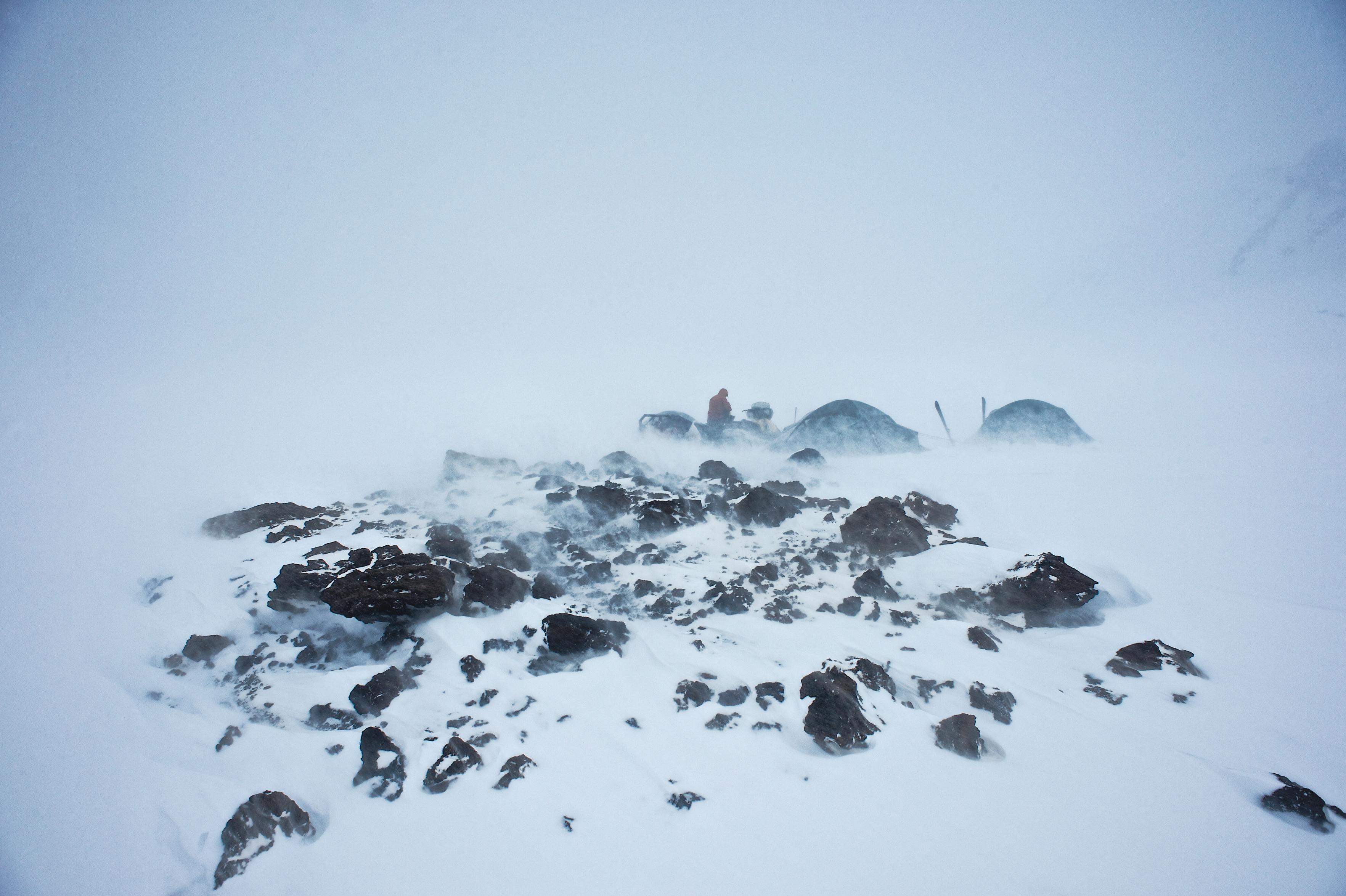 Some rocks peak through the snow in the foreground. Snow is being whipped up in the wind. Behind are the outline of some tents and a person standing