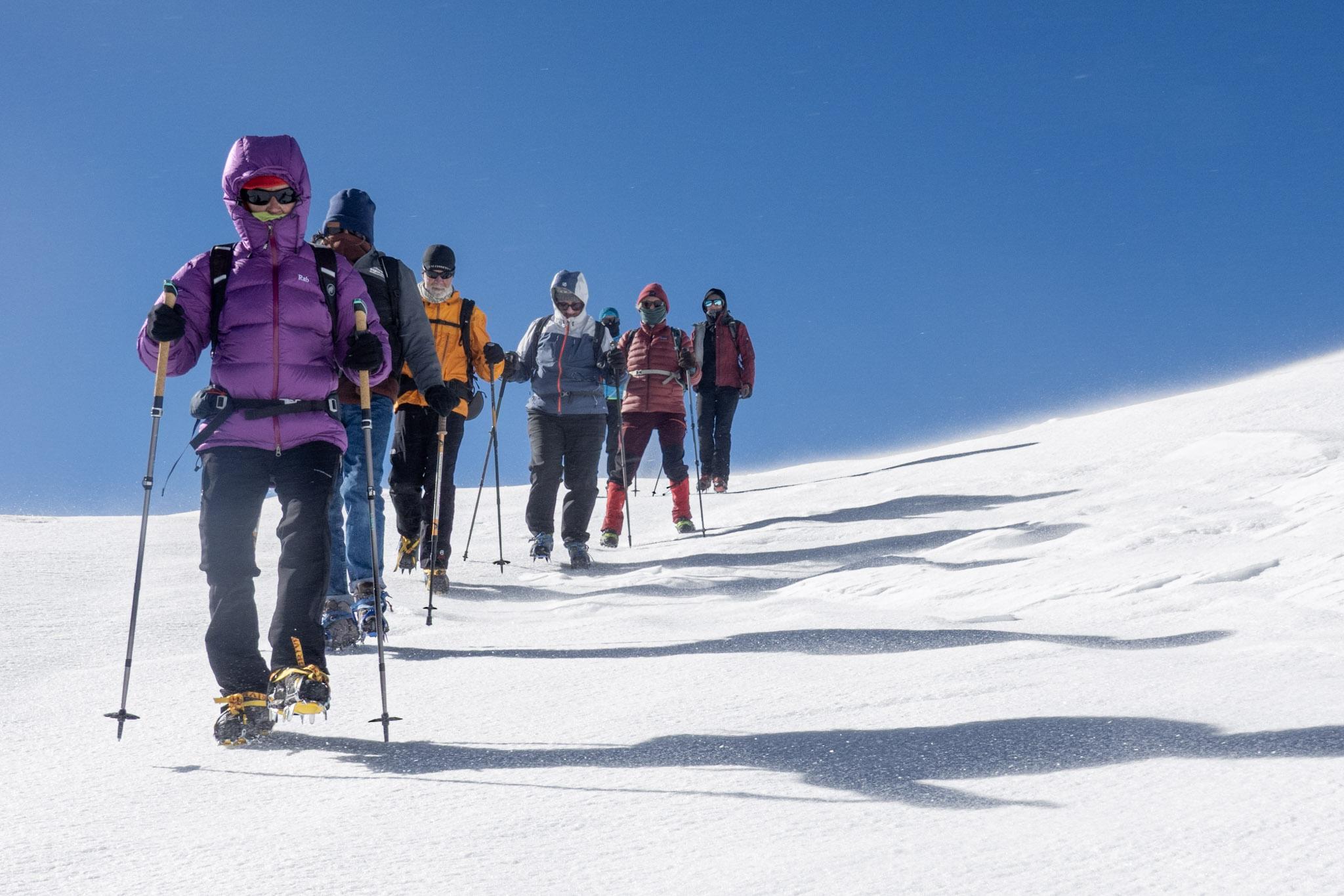 A group of colorfully dressed hikers descend a snow slope. To the right are the long shadows cast by the sun