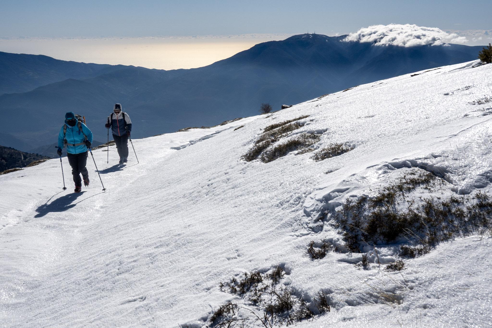 2 people pass along a snow path with iced up rocks to the right. In the background is a snow covered mountain (Sierra de Lujar) and the Mediterranean Sea