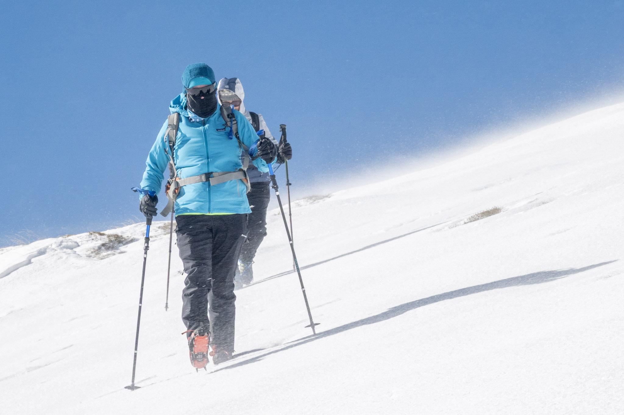 Two people descend a snow slope with wind blown spindrift
