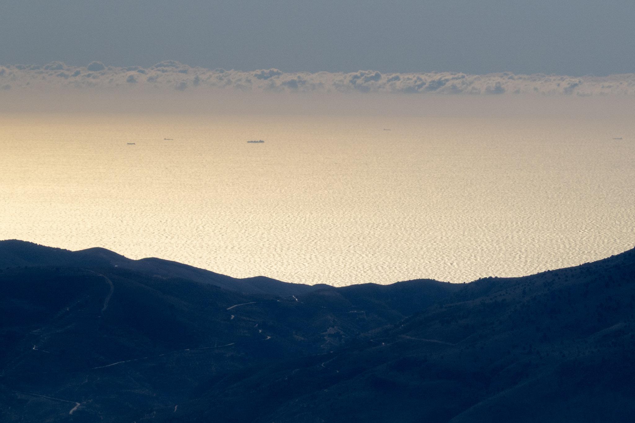 Over a small pass in the hills ships are seen on a sunlit sea. Clouds beyond