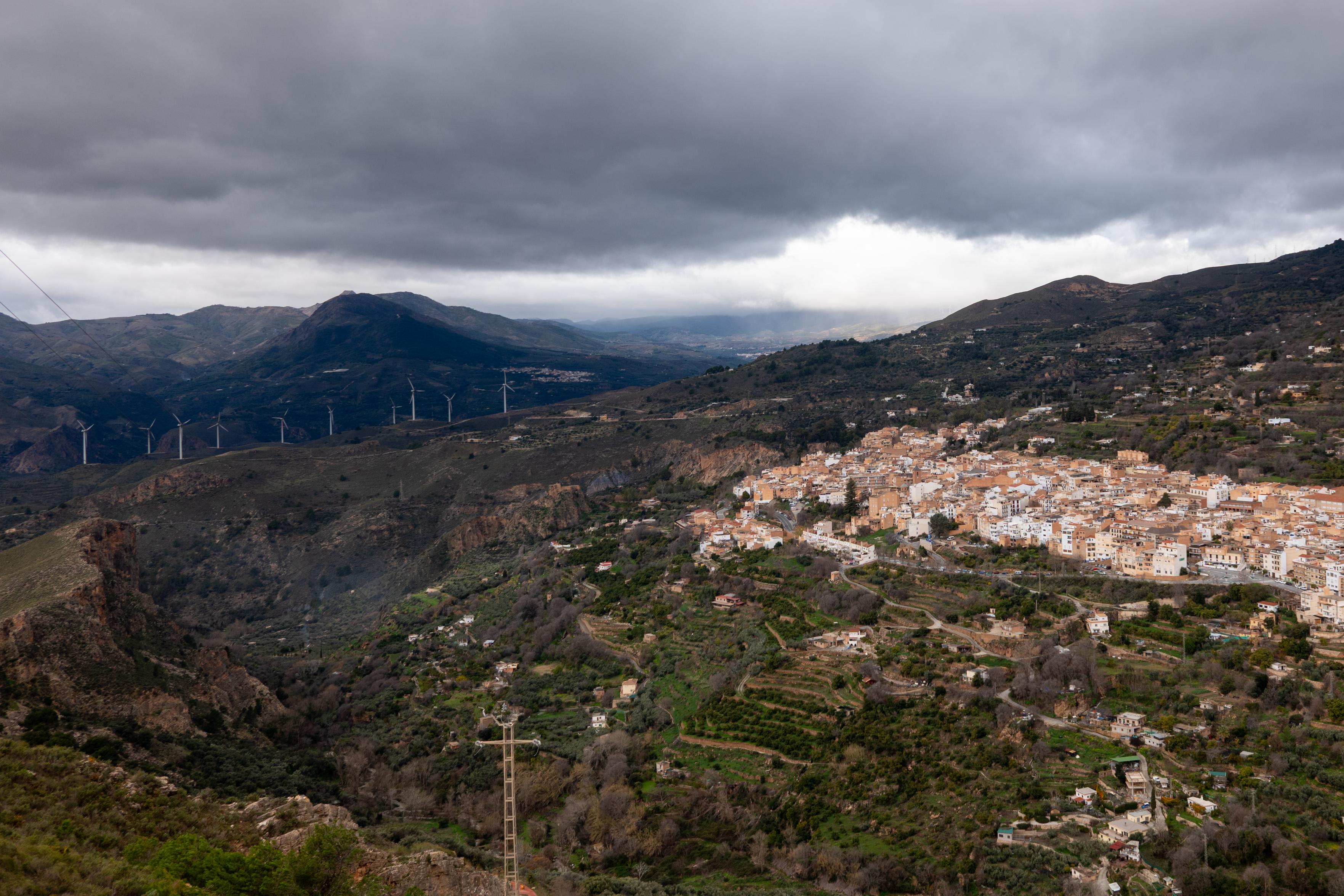 A green valley with the town of Lanjarón sat on the hillside above to the right. Behind are dark cloud and a dark mountain shape to the left (Giralda)