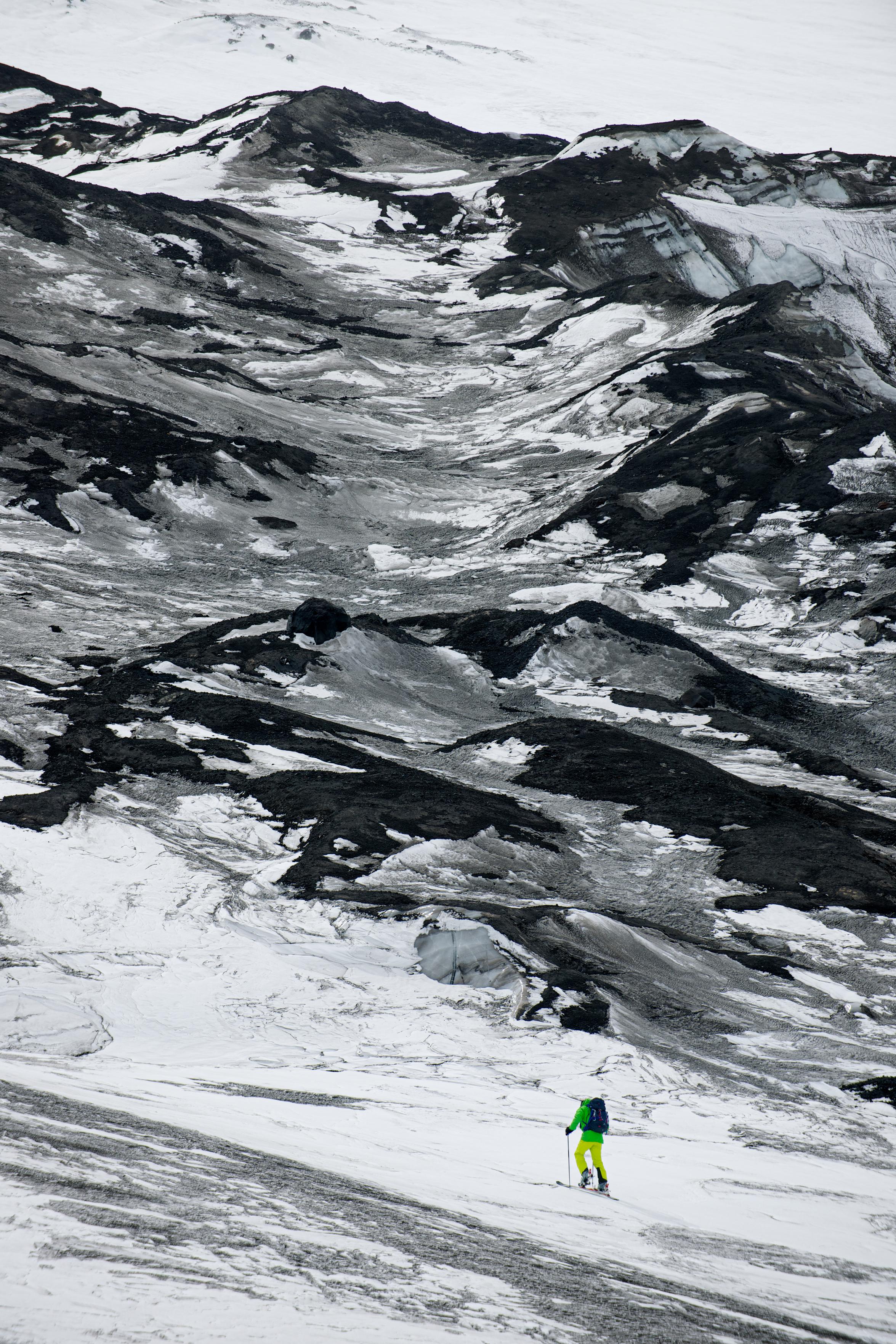 A figure in green and yellow is skiing up a white snowfield. Above and to the right are areas of bare rock, snow and volcanic ash