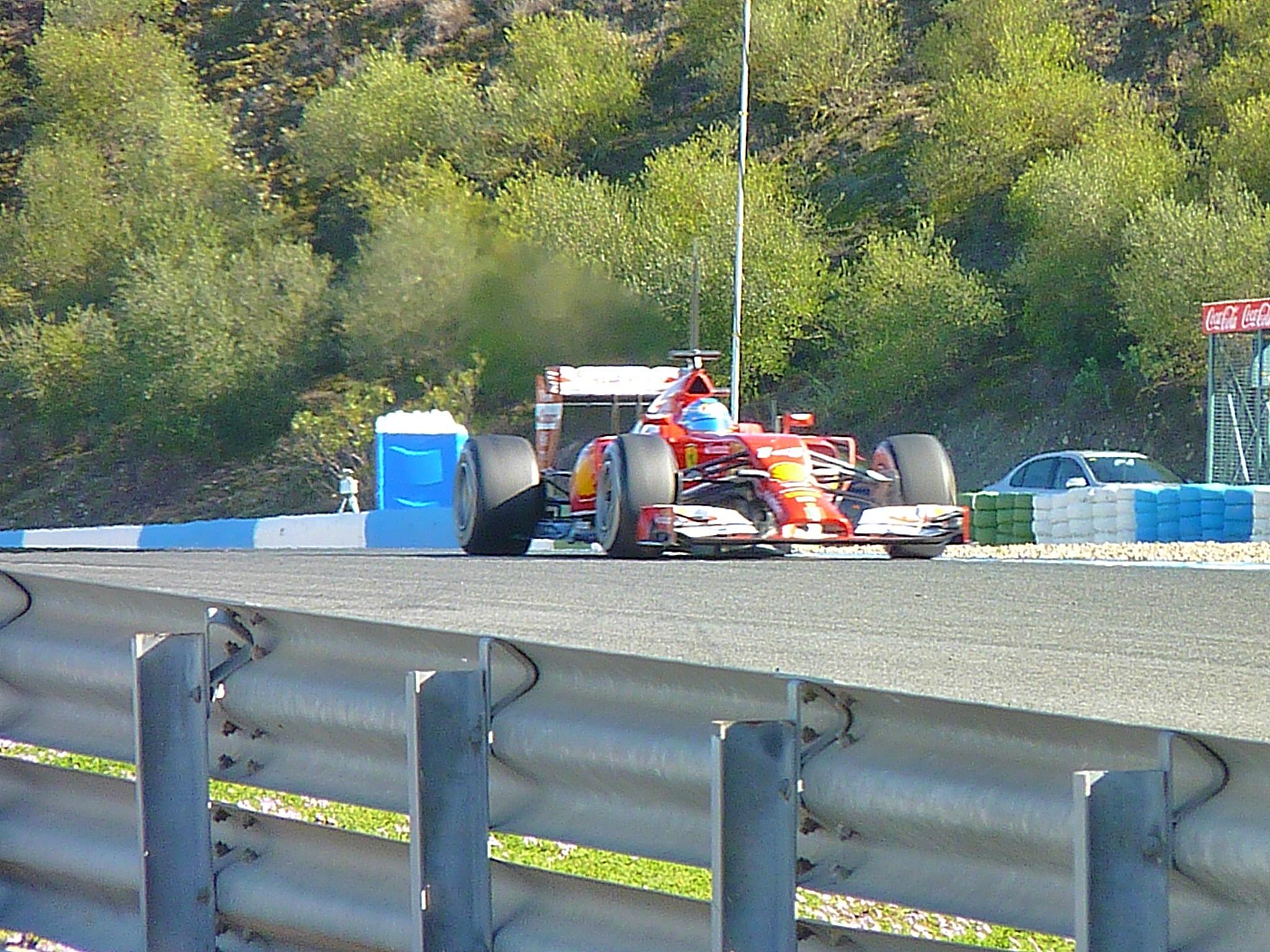 A red Ferrari F1 racing car coming down a race track. Green trees behind.