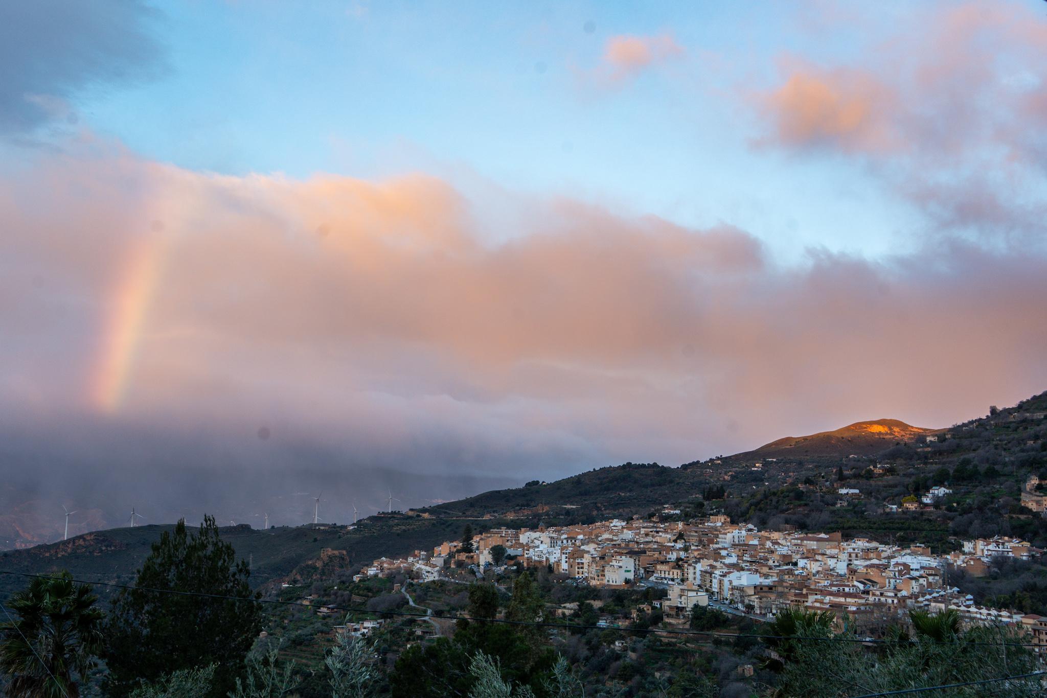Sunrise over a Spanish town. The town is in shadow apart from a small hill on the right which has sun. There are heavy clouds above and a rainbow appearing on the left