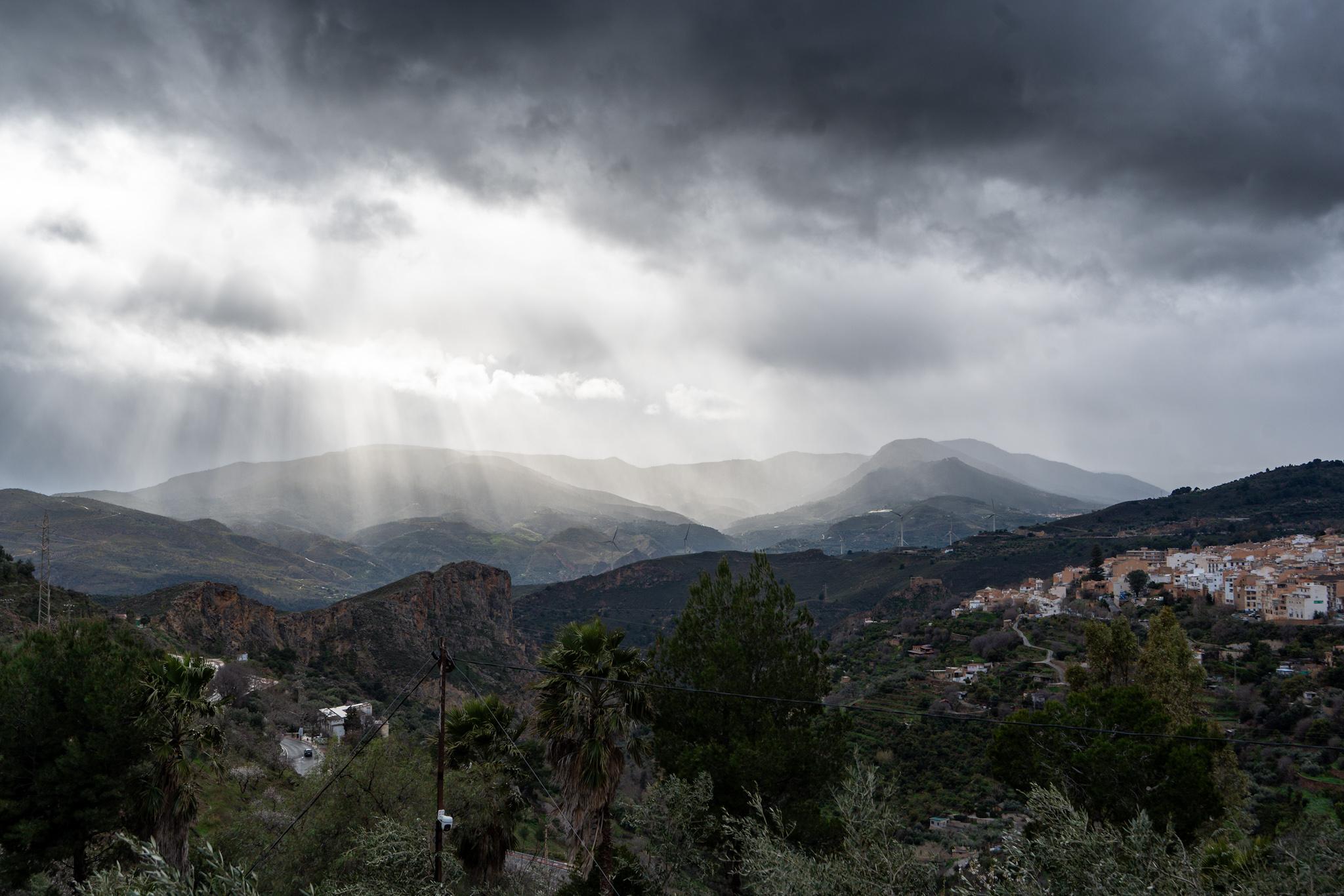 A small town lies on a plateau to the right. Below are green hills. At the back can be seen an incoming weather front with sun trying to come weakly through the dark clouds