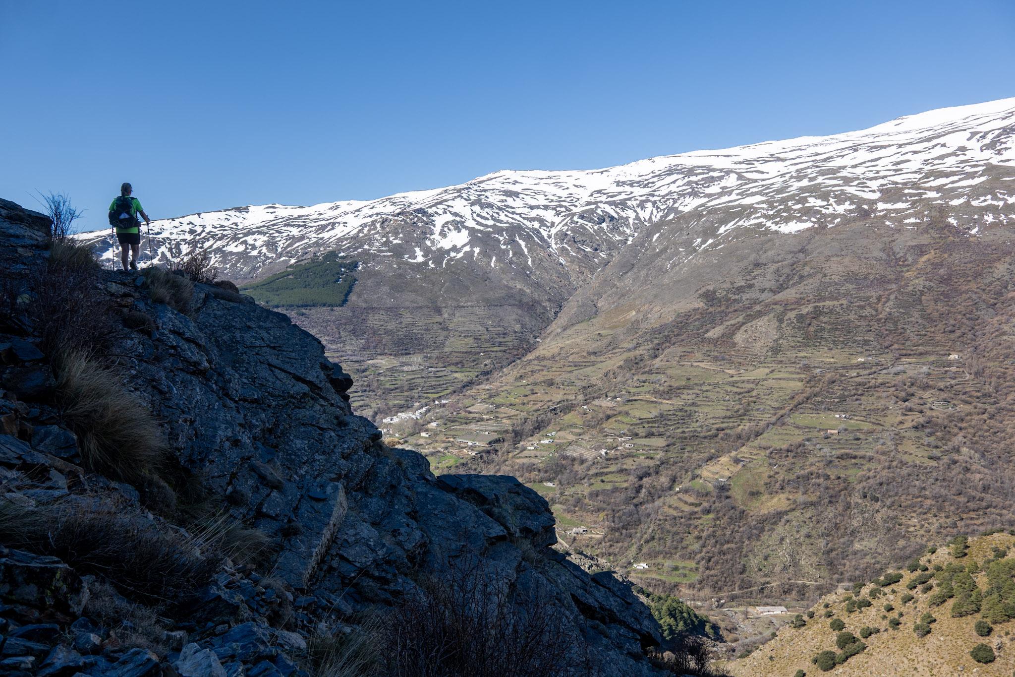 A person stands on a high trail in shadow to the left. To the right are deep valleys with snow covered mountains behind
