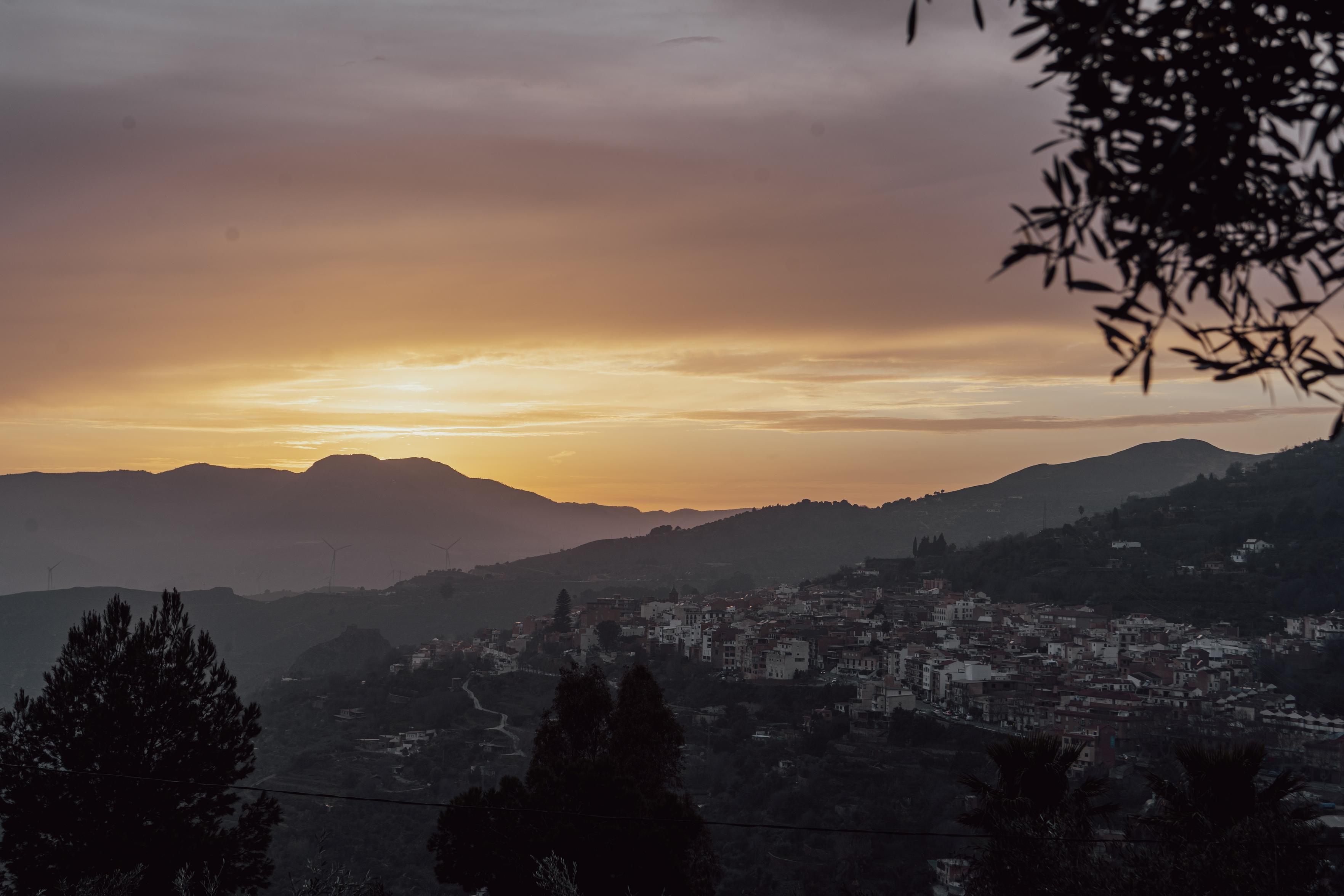 A view through some olive trees to a town at dusk surrounded by hills in the suns last light
