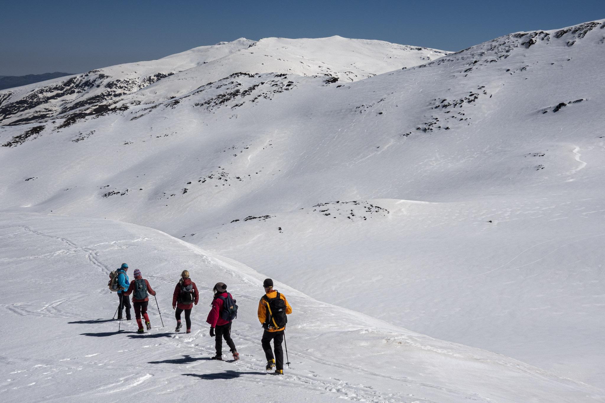 5 people hike down through the snow. Mountains in the distance