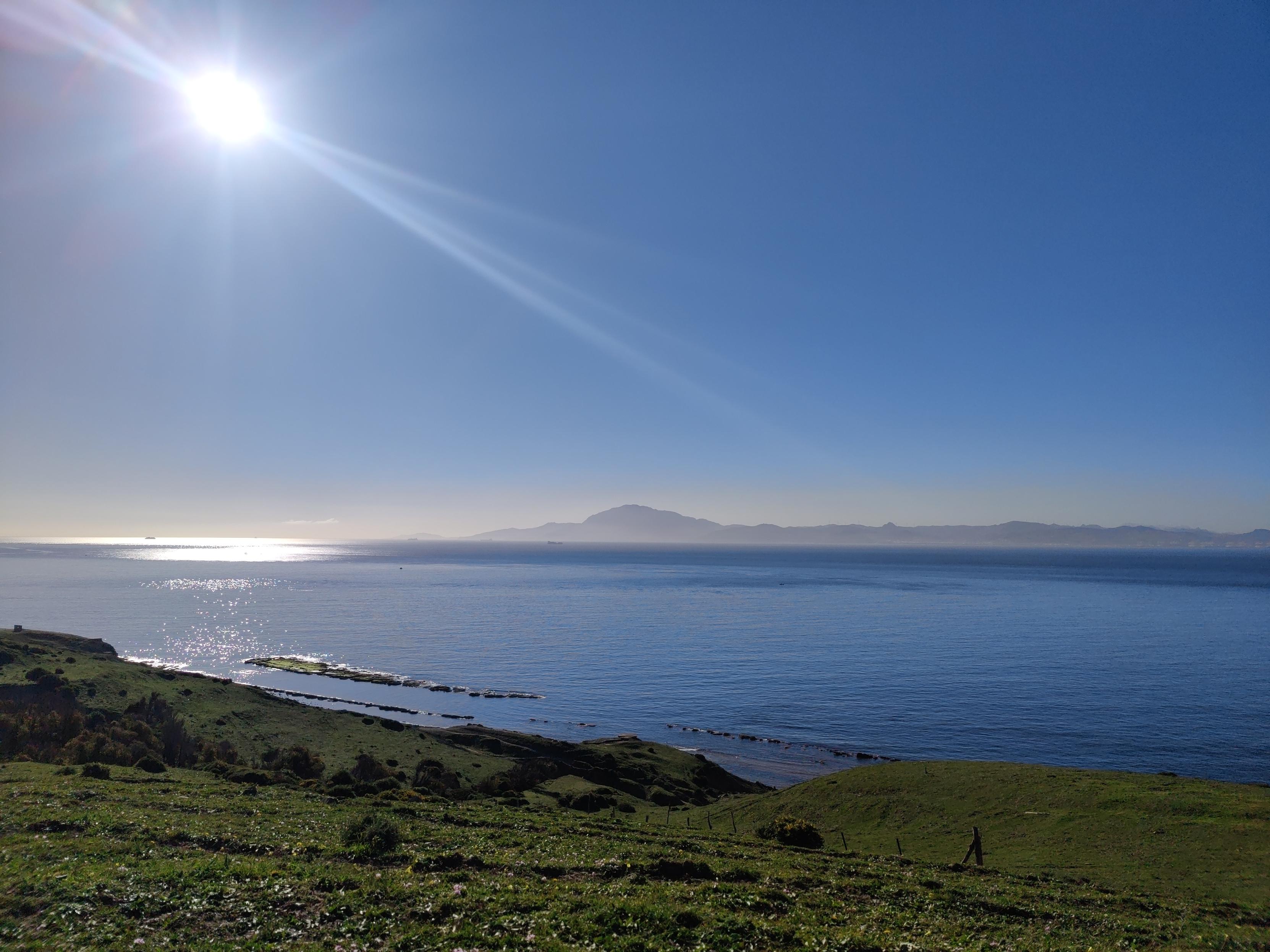 Green fields drop down to a calm sea. The sun shines on the sea in the top right. Some distant hills show in Morocco across the other side