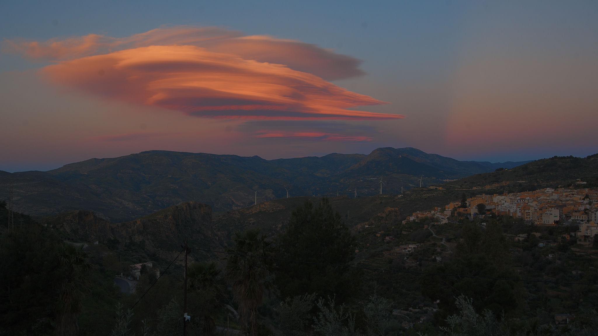 A lenticular shaped orange cloud lies above a shadowed landscape of hills and villages