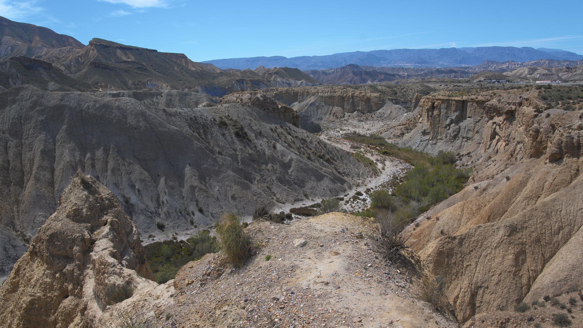 A view from a rocky headland out over a torn and twisted arid landscape of ravines and valleys. There is some signs of green vegetation in the valleys. Blue sky above