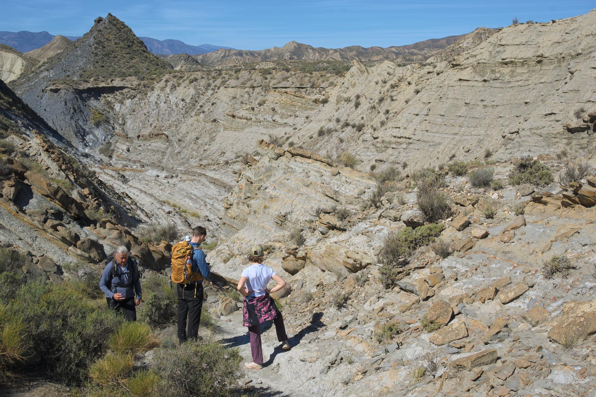 3 people stand at the entrance to an arid valley with broken rock and loose boulders
