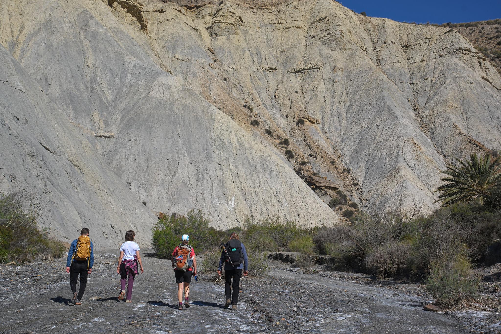 3 people hike down a wide "rambla" (valley) with sandy rock walls above and to the left and straight ahead