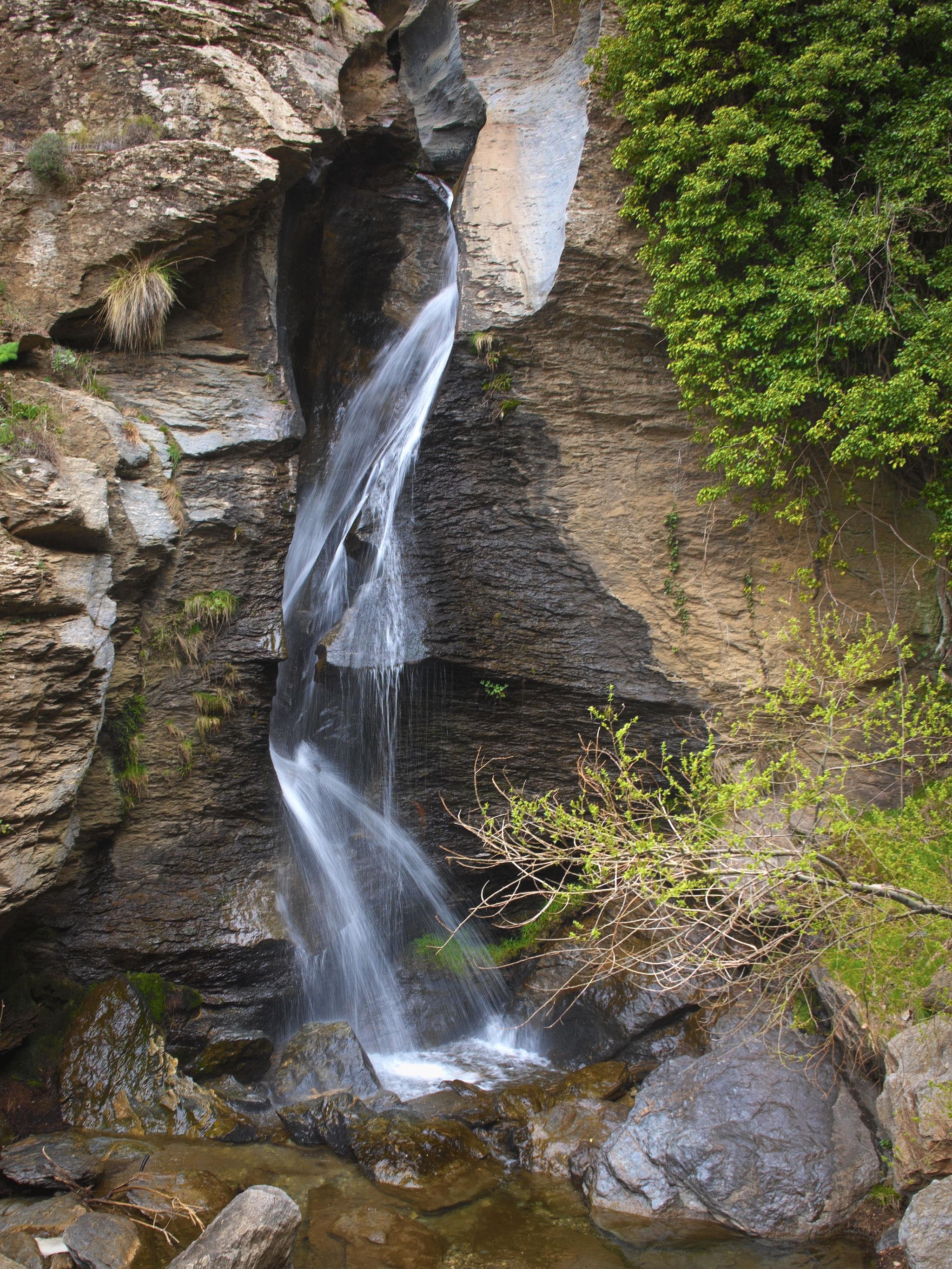 Water cascades down a narrow rift in the cliffs bouncing off both sides of the rock walls. At the bottom is a quiet pool with boulders. Lots of green trees and scrubs to the right.