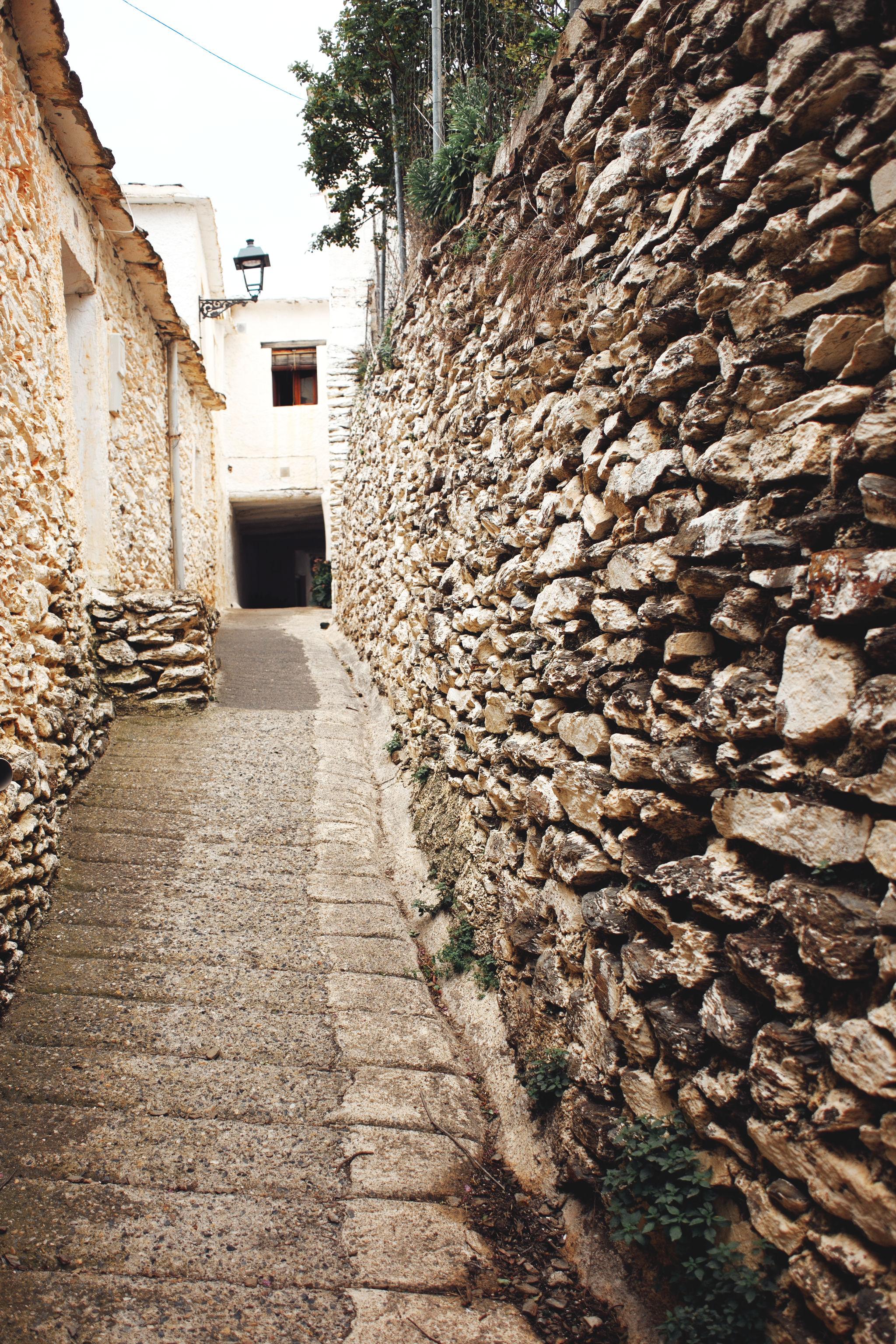 A narrow street with stone wall to the right and a white building at the end of the street