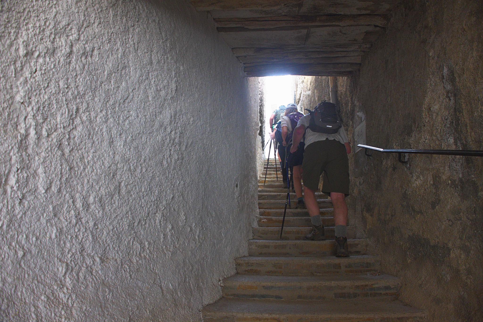 Hikers in the town of Busquistar pass up a narrow covered street. A white wall on the left and stone wall on the right
