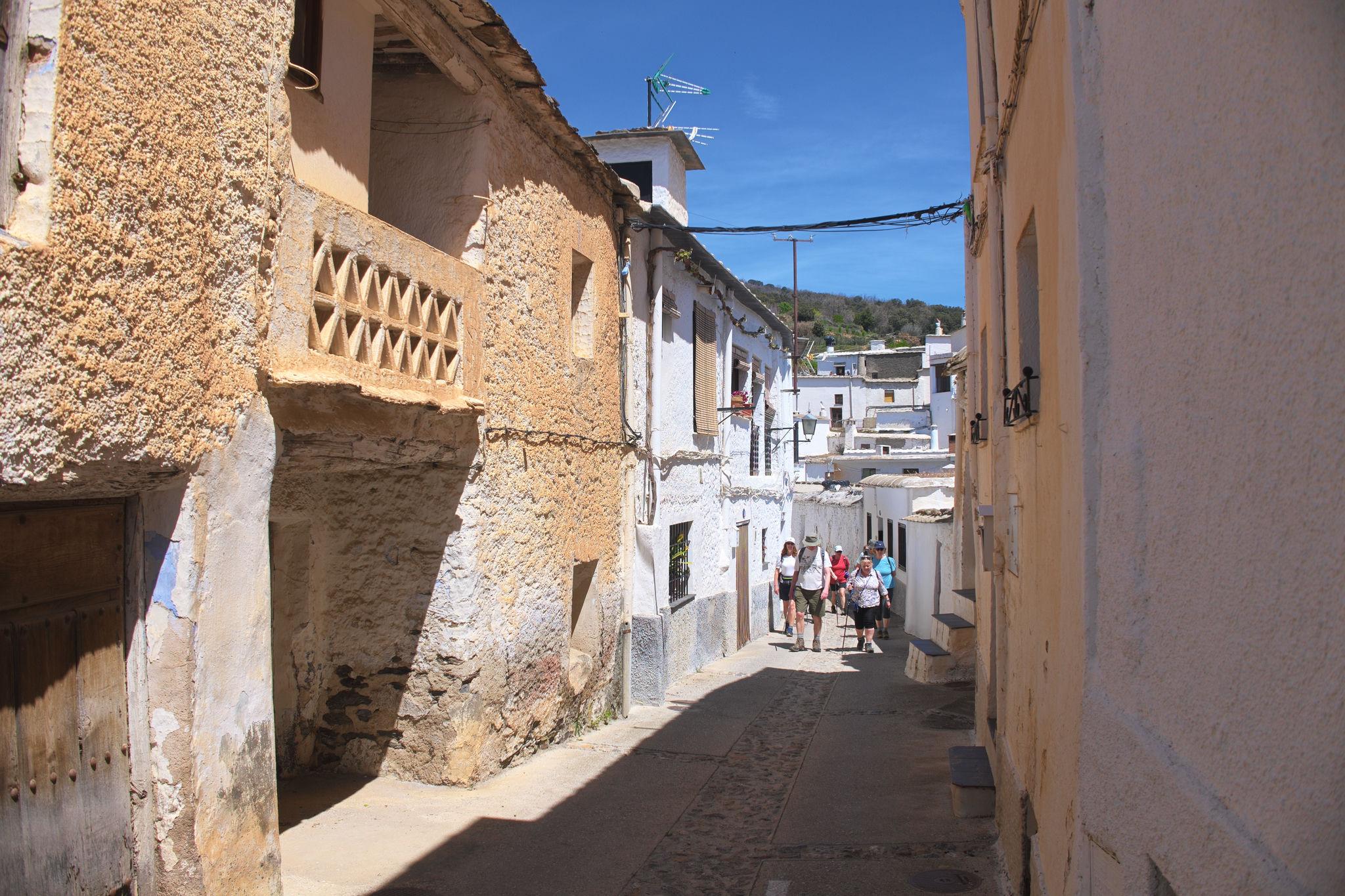Some colourful hikers pass along a street. The left hand side is in the sun and the house walls are sandy coloured with the Sahara dust storms. The right hand side is shaded