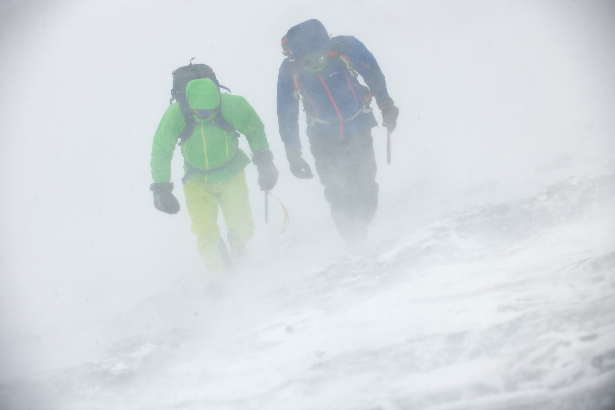 A person in green (me) leaning heavily into the wind whilst snow spindrift rushes past. Next to me is a person in red and blue jacket