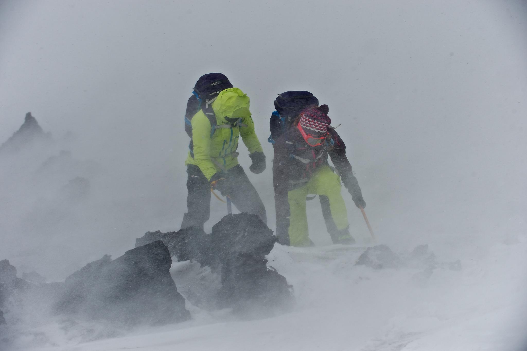 @khusky in yellow trying to walk into a very strong wind alongside Berghaus athlete, Julia Pickering. Some dark rocks appear out of the gloom