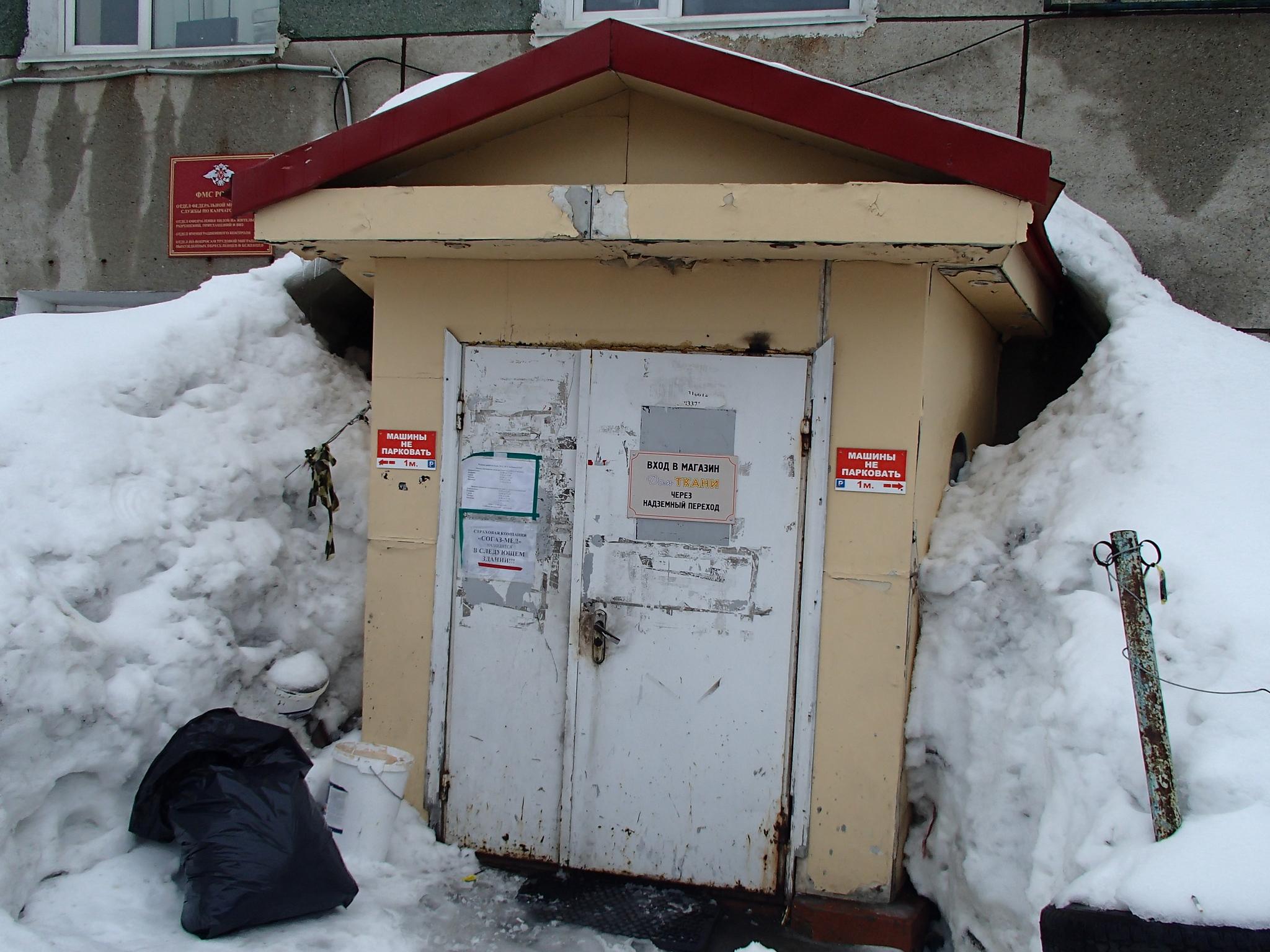 A scruffy white door with torn posters and faded writing stands surrounded by a snow drift