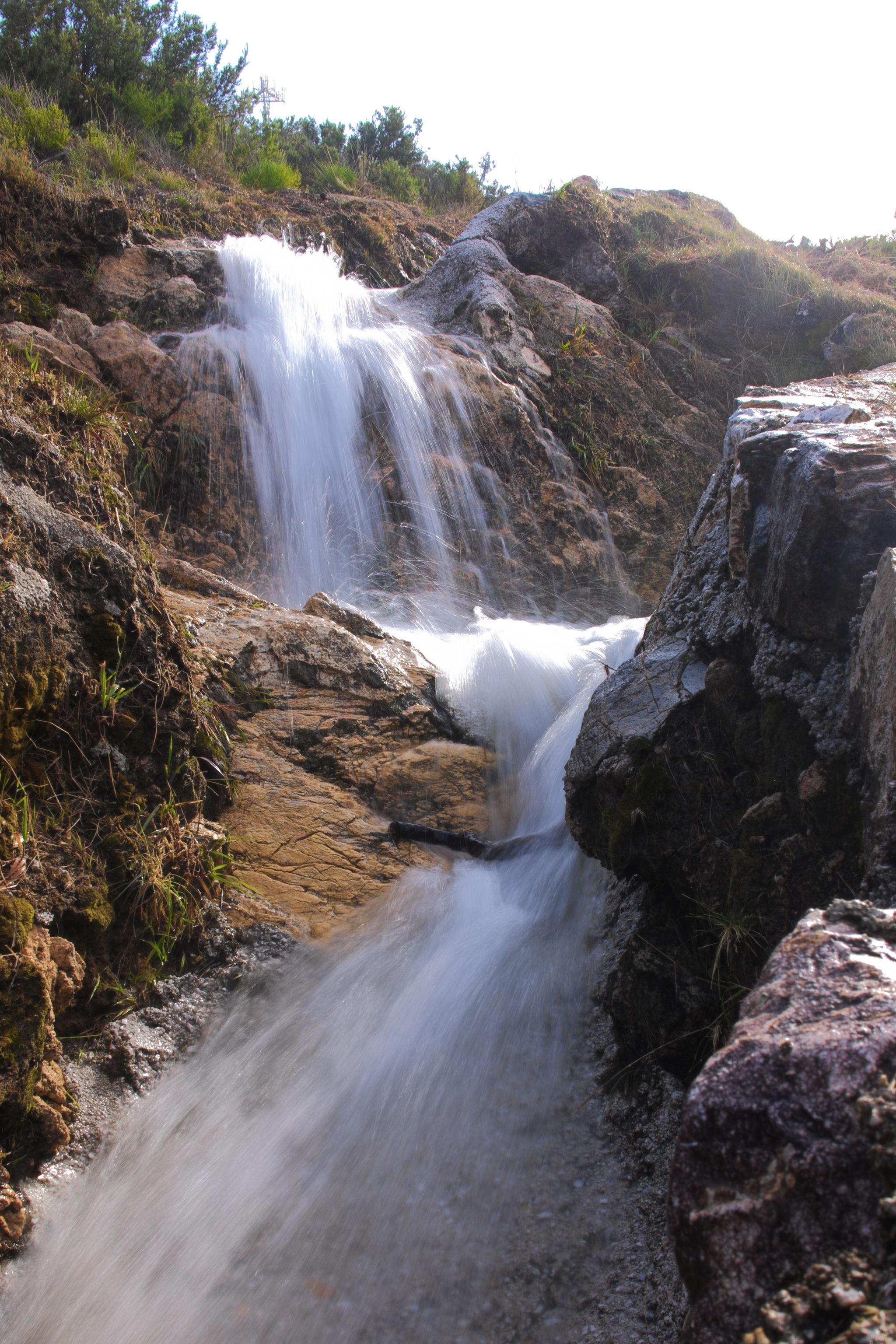 Water cascades down a series of small rock steps with rock wall to the right and trees and scrubs to the left 