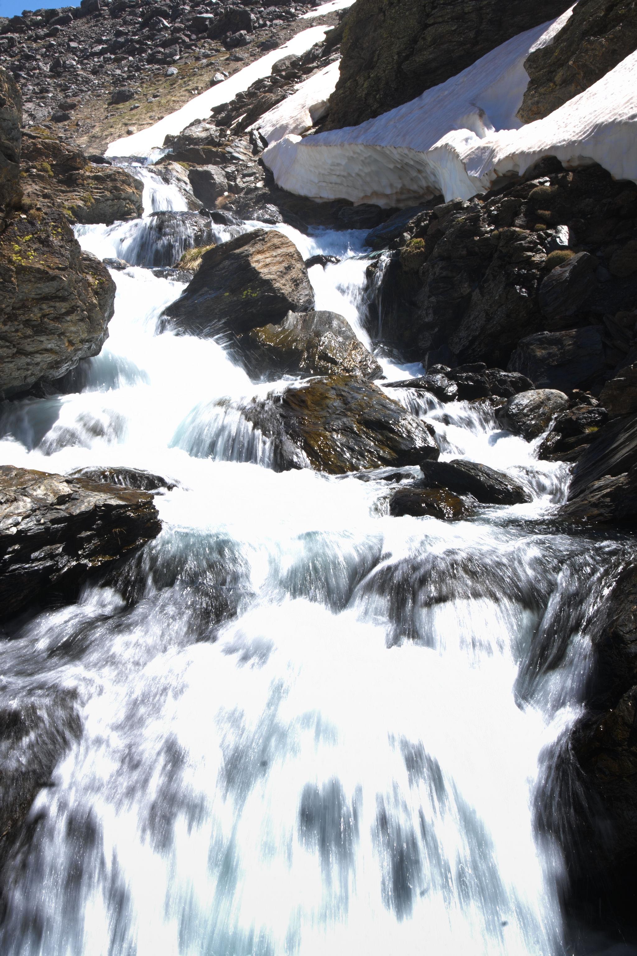 Melt water rushes down a series of waterfalls with some large snowbanks lining the right hand side of the river