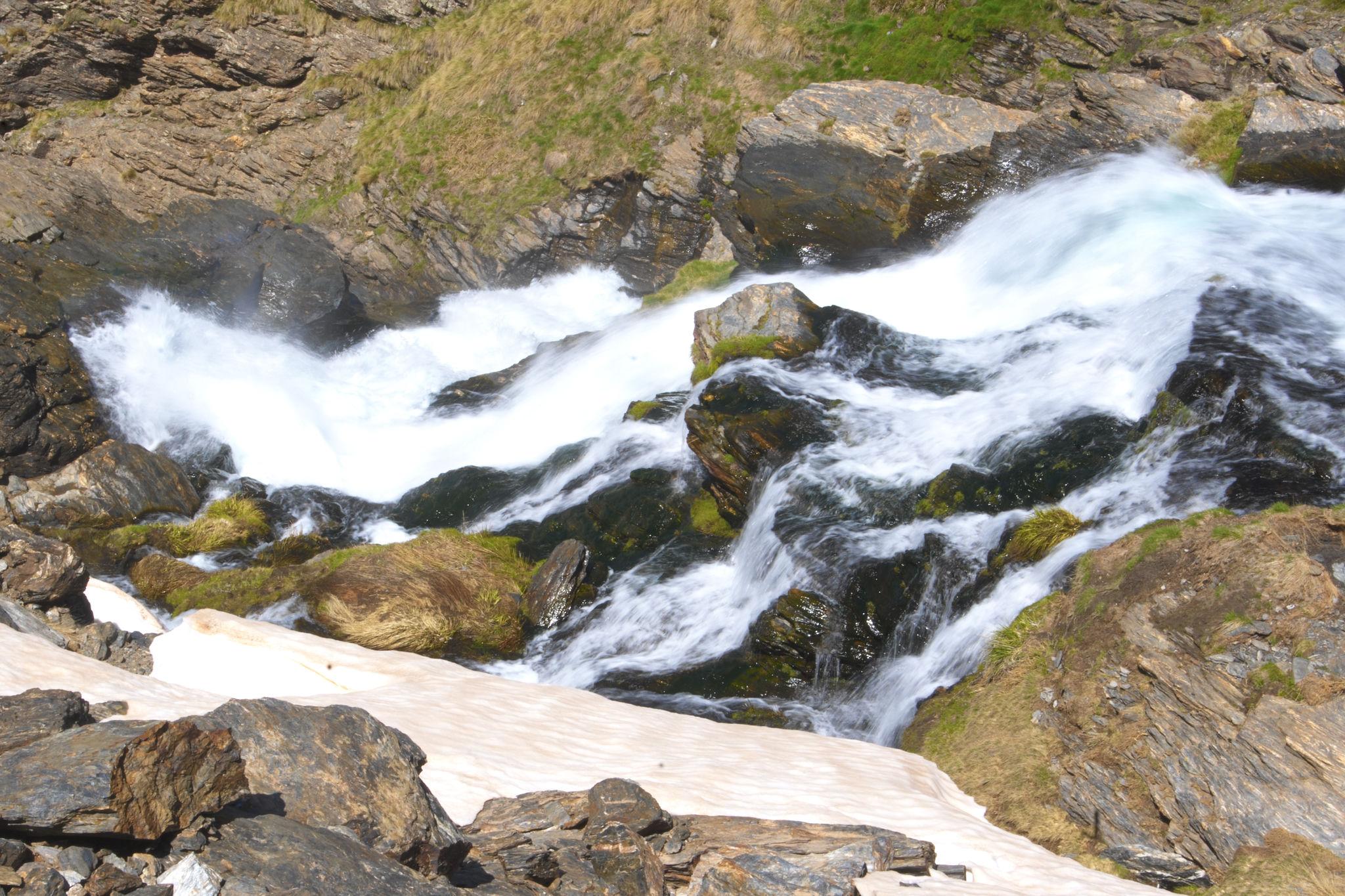 Foreground rocks emerge from a snowfield. Beyond is a river in spate crashing down a mountainside, spray thrown up into the air