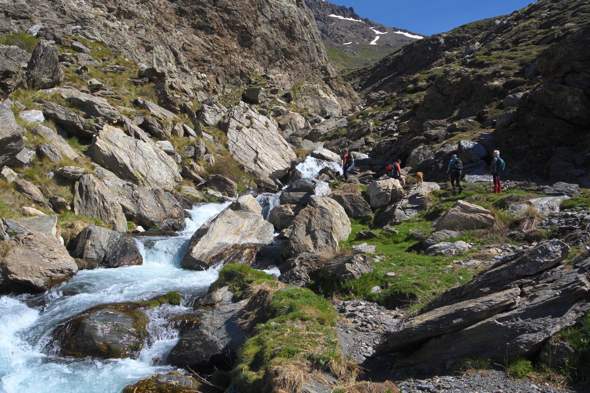 A green area to the right of a river contains some hikers. The river plunges down a series of small cascades. Mountains rise behind