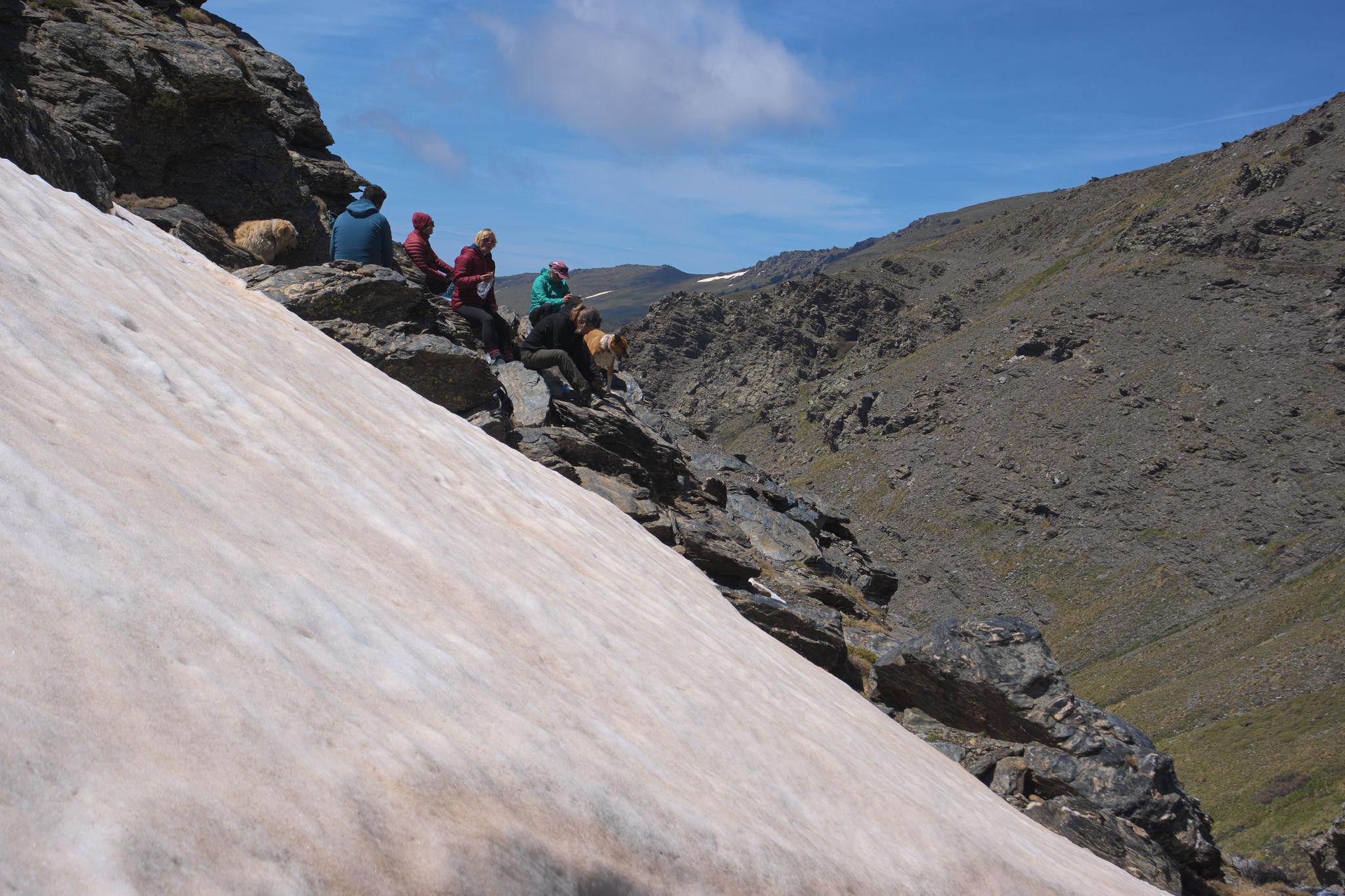 A group of hikers sit eating lunch perched on some rocks above a snow slope