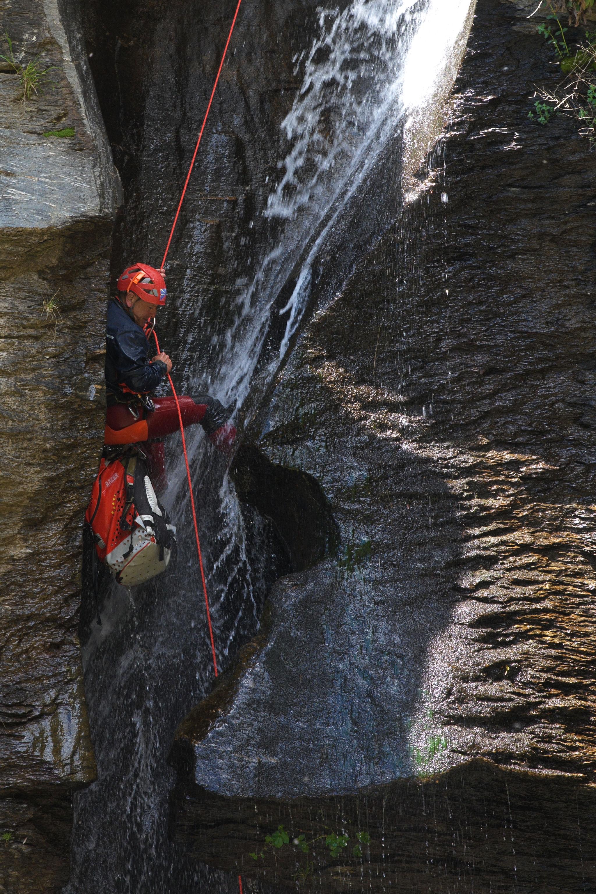 A person with orange helmet rappels down a red rope in the middle of a waterfall