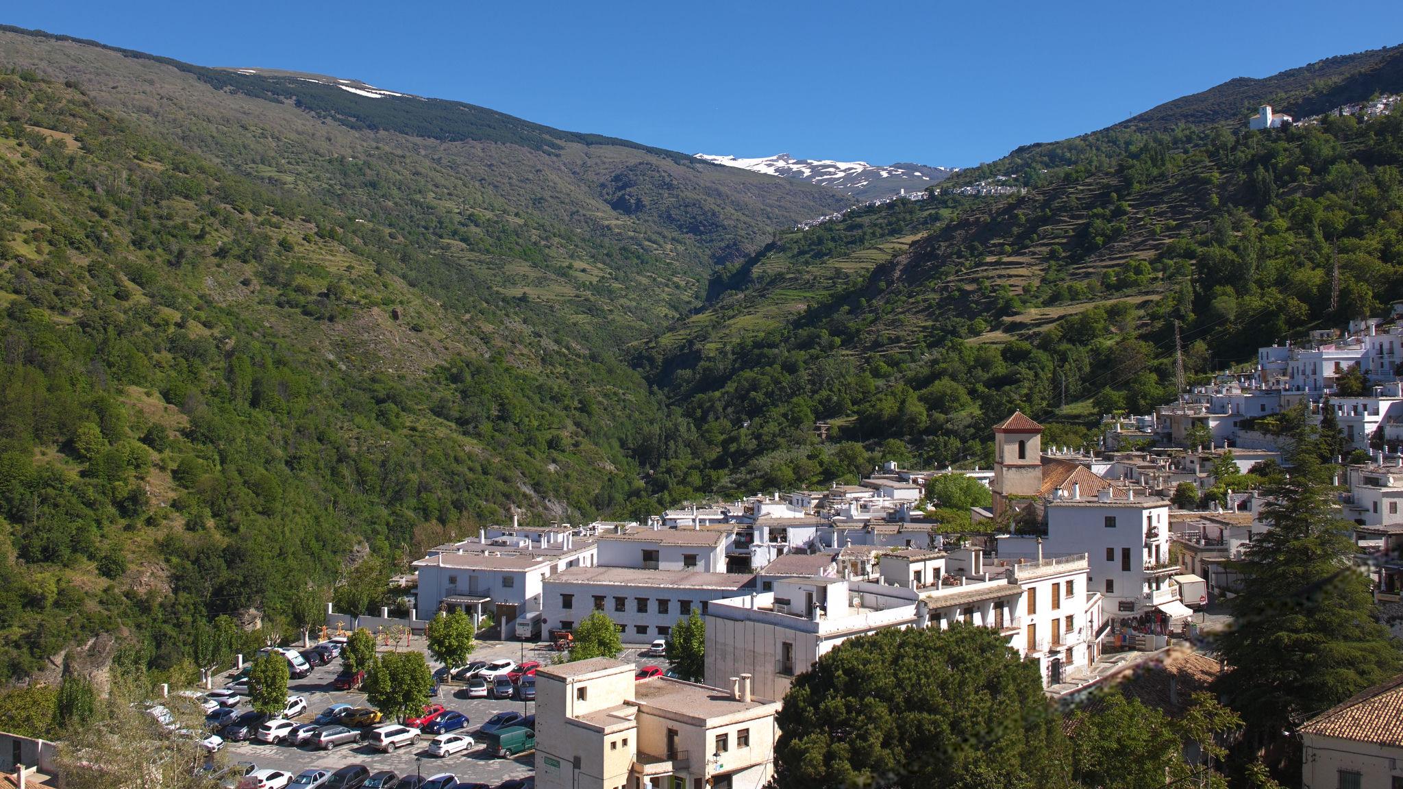 A town with white buildings sits at the foot of a long valley leading up to snow covered mountains. On the right at the head of the valley another white village can be seen